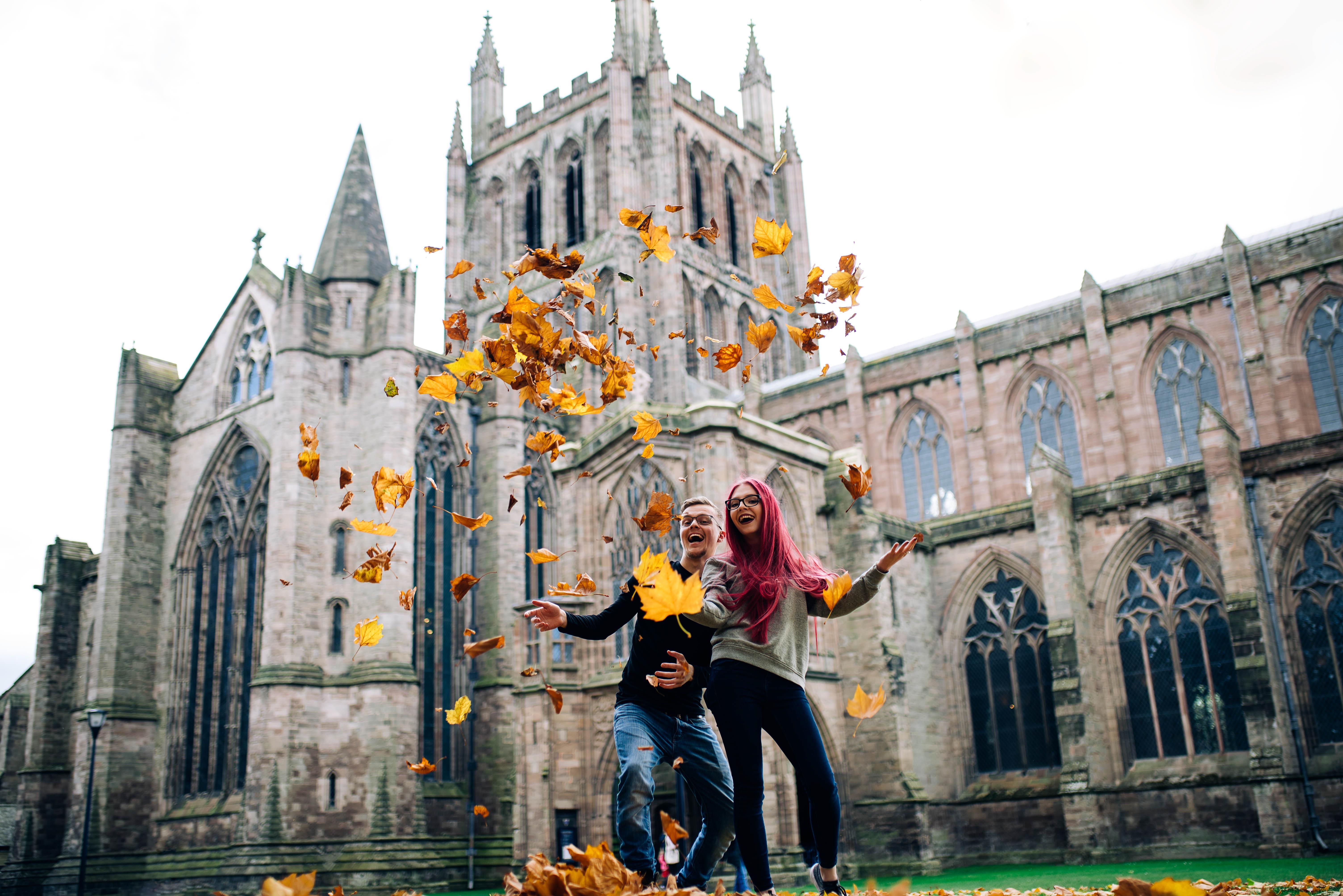 young couple smiling and throwing Autumn leaves into the air with Hereford Cathedral behind them