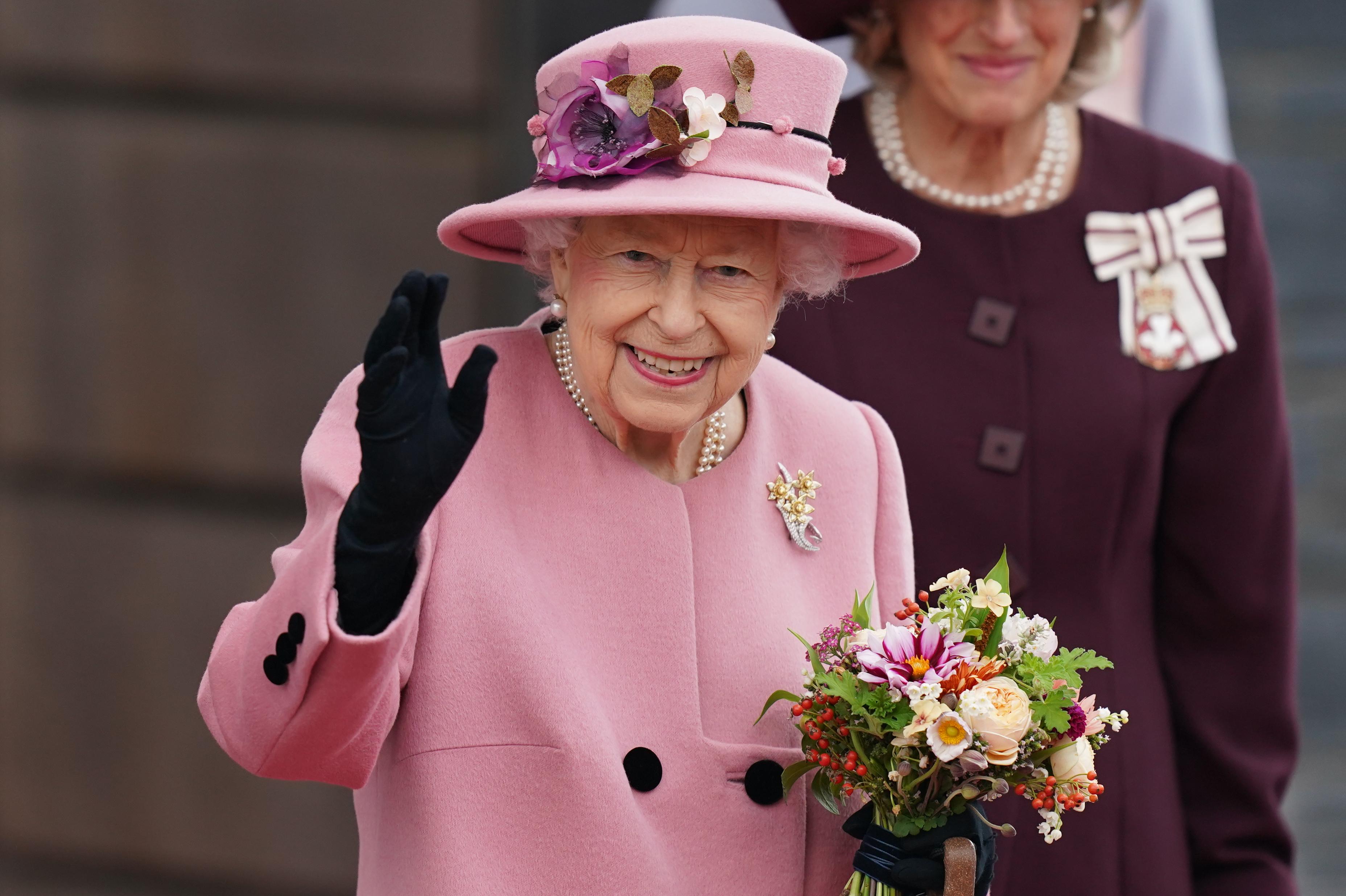 HM The Queen wearing a pink two piece suit and black gloves, smiling and waving and carrying a bouquet of flowers