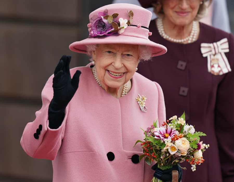 HM The Queen wearing a pink two piece suit and black gloves, smiling and waving and carrying a bouquet of flowers